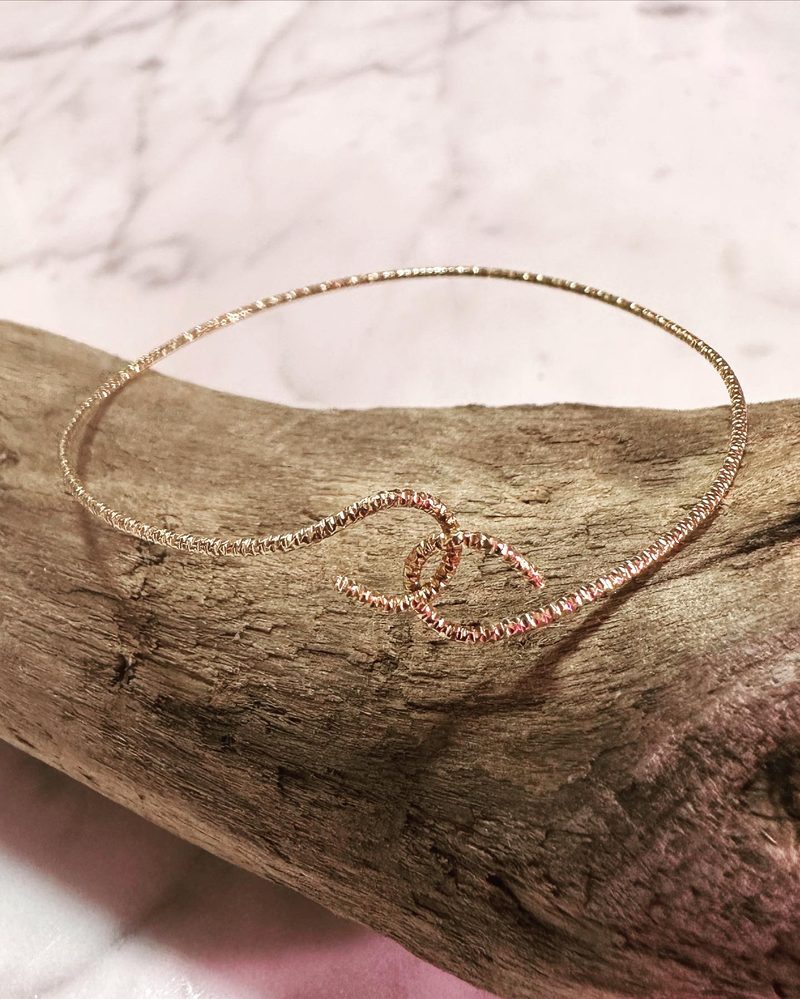 Textured gold wire bracelet with two interlocking circles at the front, resting on driftwood