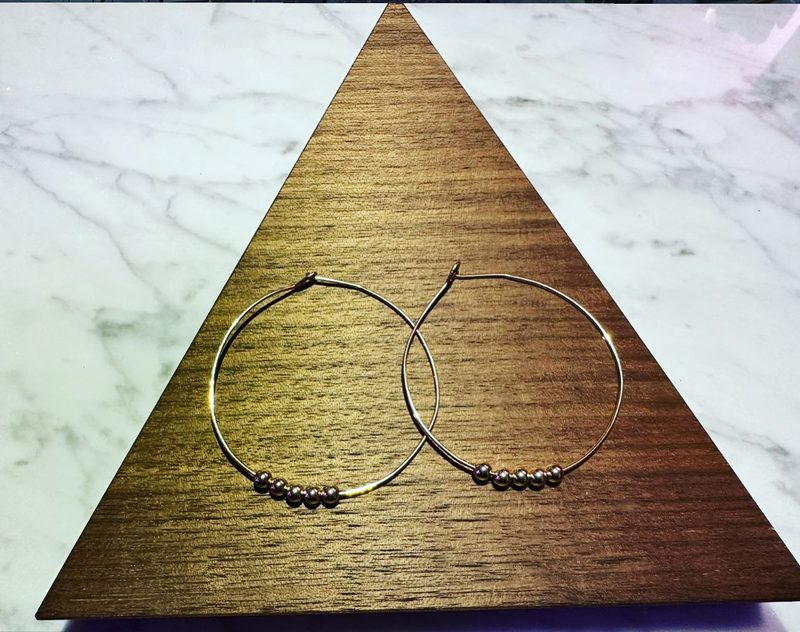 Pair of thin gold hoop earrings, each with a small cluster of dark beads, on a triangular walnut display