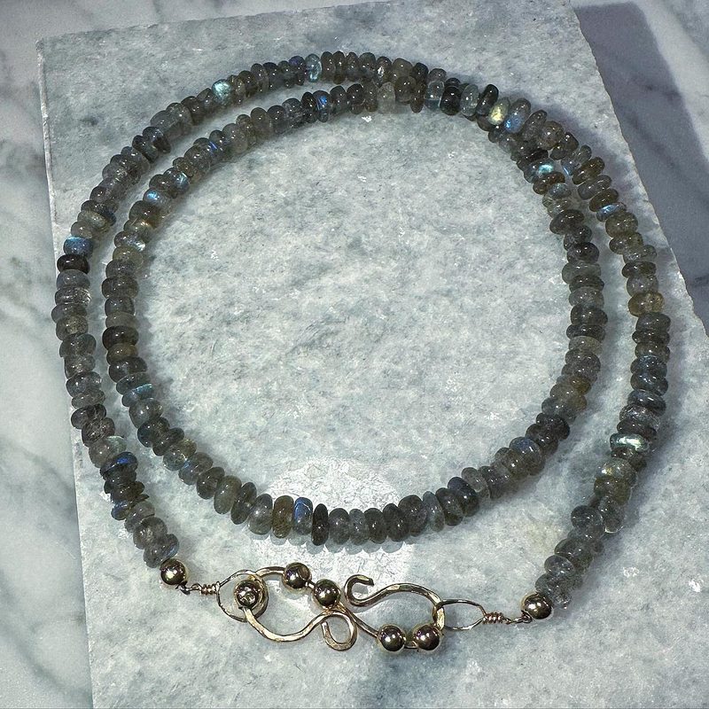 Long strand of faceted grey labradorite beads with a gold-filled hook clasp, laid out on white marble